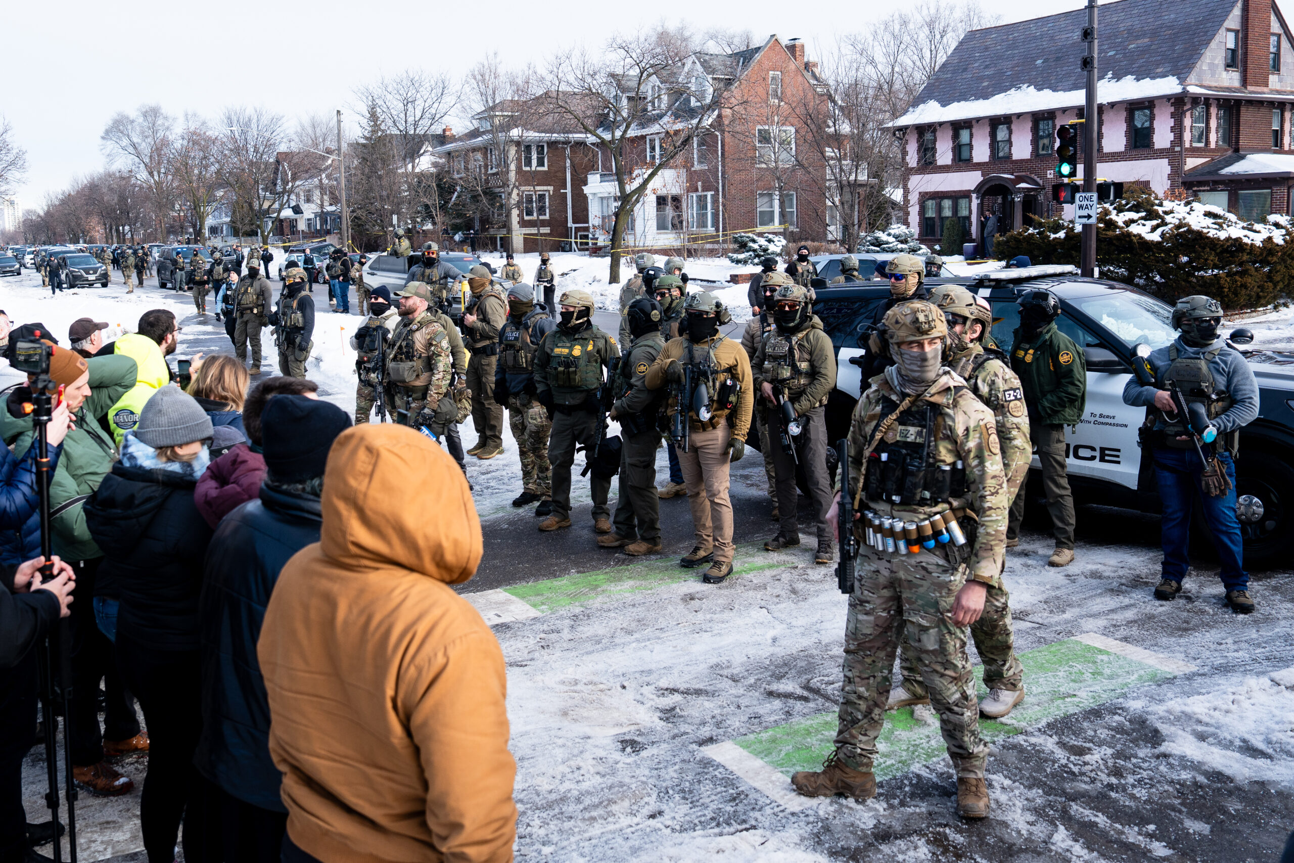 Armed ICE agents stand on a snowy Minneapolis street as residents watch from the sidewalk.